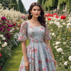 Puede incluir: Una mujer lleva un vestido floral azul claro con escote cuadrado y mangas cortas abullonadas. El vestido tiene un corpiño ajustado y una falda fluida. Está de pie en un jardín con flores en el fondo.