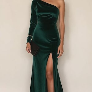 K&ouml;nnte beinhalten: Ein langes, dunkelgr&uuml;nes Samtkleid mit einem einseitigen Ausschnitt und einem hohen Schlitz am Oberschenkel. Das Kleid hat ein figurbetontes Oberteil und einen flie&szlig;enden Rock.