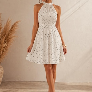 K&ouml;nnte beinhalten: Wei&szlig;es Halfterkleid mit schwarzen Polka Dots. Das Kleid hat eine taillierte Taille und einen ausgestellten Rock. Das Model tr&auml;gt Riemchenabs&auml;tze. Ein klassisches und vielseitiges Kleidungsst&uuml;ck.