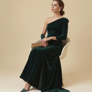 K&ouml;nnte beinhalten: Eine Frau in einem dunkelgr&uuml;nen Samtkleid mit einer Schulter, die auf einem wei&szlig;en Stuhl sitzt. Sie h&auml;lt eine braune Clutch und schaut zur Seite.