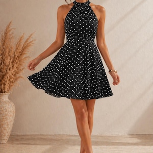 K&ouml;nnte beinhalten: Schwarzes Neckholder-Kleid mit wei&szlig;en Polka-Dots. Das Kleid hat ein figurbetontes Oberteil und einen ausgestellten Rock. Das Model tr&auml;gt beige Abs&auml;tze und ein goldenes Armband. Der Hintergrund ist neutral.