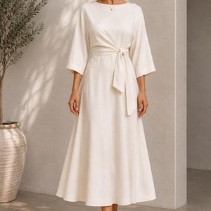 May include: A cream-colored midi dress featuring a round neckline, three-quarter length sleeves, and a wrap-style waist with a matching fabric tie belt. The skirt flows into a wide, A-line silhouette that falls below the knee in a solid color.