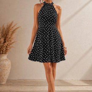 K&ouml;nnte beinhalten: Ein schwarzes Kleid mit wei&szlig;en Punkten und einem Neckholder-Ausschnitt. Das Kleid hat ein figurbetontes Oberteil und einen ausgestellten Rock. Das Model tr&auml;gt beige Abs&auml;tze. Der Hintergrund ist neutral.