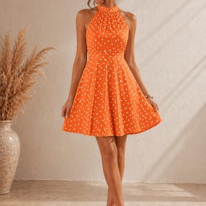 K&ouml;nnte beinhalten: Ein orangefarbenes Neckholder-Kleid mit wei&szlig;em Polka-Dot-Muster. Das Kleid hat ein figurbetontes Oberteil und einen ausgestellten Rock. Das Model tr&auml;gt beige Abs&auml;tze. Das Kleid ist ein Sommer- oder Fr&uuml;hlingsstil.