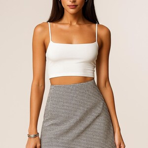 K&ouml;nnte beinhalten: Eine Frau tr&auml;gt ein wei&szlig;es Crop-Top mit d&uuml;nnen Tr&auml;gern und einen hoch taillierten Minirock mit Hahnentrittmuster. Der Rock hat ein schwarz-wei&szlig;es Karomuster. Sie tr&auml;gt auch ein silbernes Armband.
