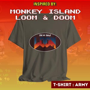 Könnte beinhalten: Armeegrünes T-Shirt mit dem Text "ASK ME ABOUT LOOM" in einem Oval. Das Wort "LOOM" ist in einer stilisierten Schriftart mit blauen, orangefarbenen und roten Farben gehalten. Auf der Oberseite des Shirts steht "INSPIRED BY: MONKEY ISLAND LOOM & DOOM".
