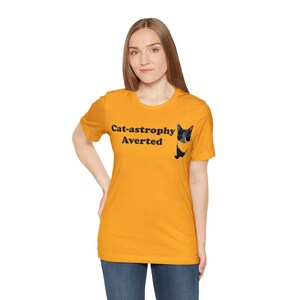 May include: A yellow t-shirt with the text "Cat-astrophy Averted" and a black and white cat peeking out from behind the text.