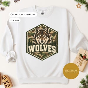 May include: A white crewneck sweatshirt featuring a central graphic of a wolf head inside a hexagon. The hexagon has a camouflage pattern and a forest silhouette. The word WOLVES is printed in large, bold, light-colored capital letters below the wolf.