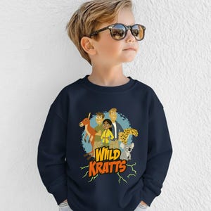 May include: Navy blue sweatshirt featuring a colorful graphic of characters and animals from the animated series "Wild Kratts." The design includes a kangaroo, cheetah, and koala. The words "Wild Kratts" are in orange and yellow.