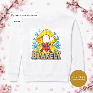 May include: A white sweatshirt featuring a graphic of a yellow hooded raincoat with a red number 4 and the name BLAKELY in stone-textured letters, surrounded by blue water droplets and small green grass patches.