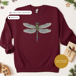 May include: A maroon long-sleeve crewneck sweatshirt featuring a detailed graphic of a dragonfly with iridescent green and blue wings and body centered on the chest. The garment has a ribbed collar, cuffs, and hem, with a standard fit.
