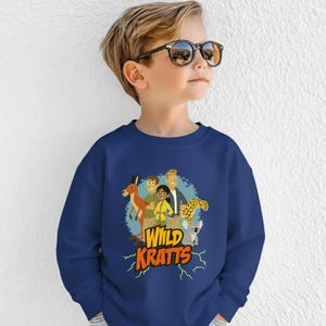 May include: A navy blue sweatshirt featuring a colorful graphic of cartoon characters and animals, including a kangaroo, cheetah, and koala. The text "Wild Kratts" is displayed in yellow with a lightning bolt design.
