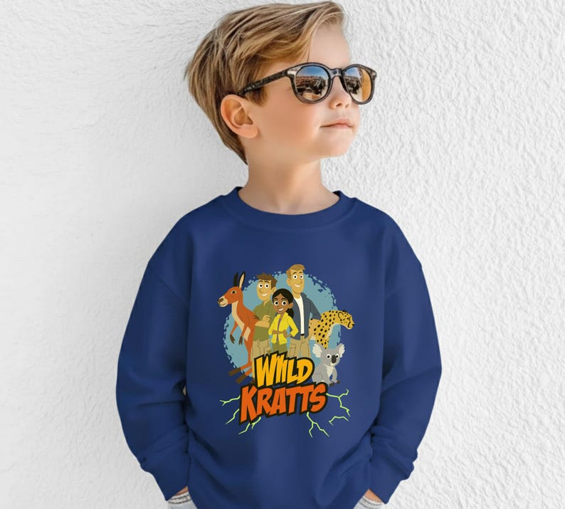 May include: A navy blue sweatshirt featuring a colorful graphic of cartoon characters and animals, including a kangaroo, cheetah, and koala. The text "Wild Kratts" is displayed in yellow with a lightning bolt design.