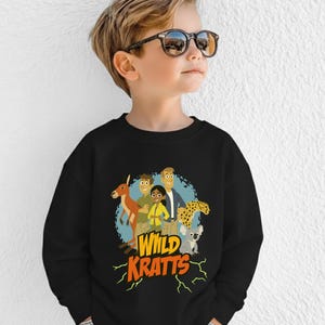 May include: Black sweatshirt featuring a colorful graphic of cartoon characters and animals, including a kangaroo, cheetah, and koala. The text "Wild Kratts" is displayed in yellow and orange. The sweatshirt is worn by a child.
