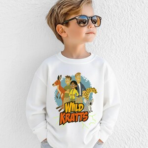 May include: White sweatshirt featuring a colorful graphic of the Wild Kratts characters with a kangaroo, cheetah, and koala. The text "Wild Kratts" is in yellow and orange. The sweatshirt is worn by a child.