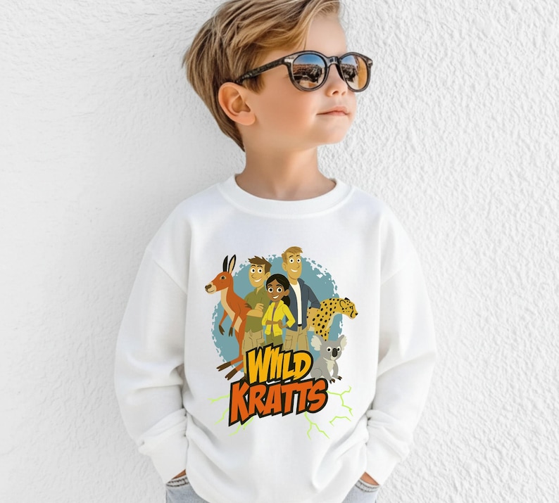 May include: White sweatshirt featuring a colorful graphic of the Wild Kratts characters with a kangaroo, cheetah, and koala. The text "Wild Kratts" is in yellow and orange. The sweatshirt is worn by a child.
