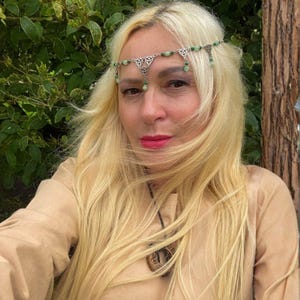 May include: A silver-toned headpiece with green beads and Celtic knot designs. The headpiece is worn on the forehead. The person in the image is wearing a tan-colored top and has long blonde hair.