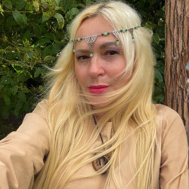 May include: A silver-toned headpiece with green beads and Celtic knot designs. The headpiece is worn on the forehead. The person in the image is wearing a tan-colored top and has long blonde hair.