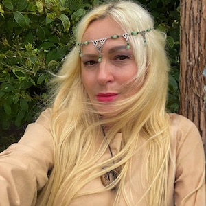 May include: A silver-toned headpiece featuring intricate metalwork and small green beads, worn across the forehead. The accessory includes triangular filigree elements and dangling green accents, complemented by a dark cord necklace with a circular pendant.