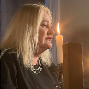 May include: A woman with blonde hair wearing a black top and a silver necklace. A lit white candle is in the background, casting a warm glow. The candle is in a wooden candle holder.