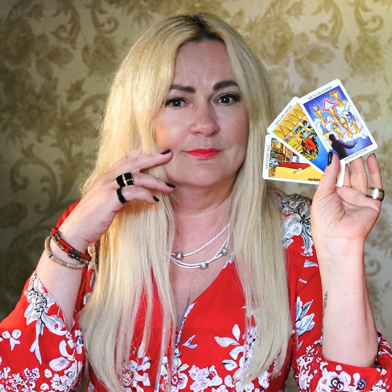 以下が含まれることがあります： 手元に持たれた3枚のタロットカード。魔術師、カップの8、カップの7が描かれています。カードにはカラフルなイラストとテキストラベルがあります。人物は赤い花柄のブラウスを着用し、ネックレスと指輪を身につけています。