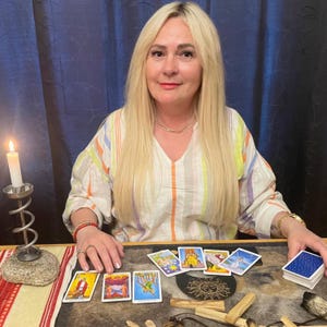 May include: A woman with long blonde hair sits at a table with tarot cards. A lit candle in a metal holder is to the left. Several tarot cards are laid out, and a deck of cards is on the right. The woman wears a striped shirt.