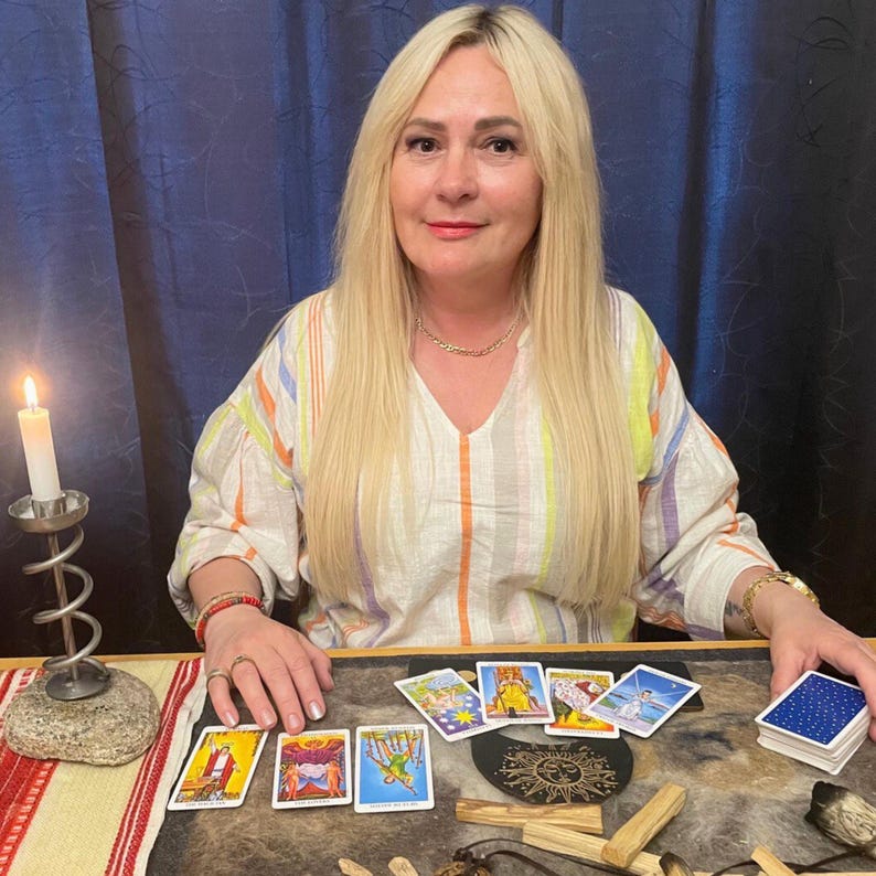 May include: A woman with long blonde hair sits at a table with tarot cards. A lit candle in a metal holder is to the left. Several tarot cards are laid out, and a deck of cards is on the right. The woman wears a striped shirt.