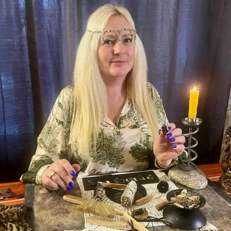 May include: A woman with long blonde hair wearing a green and white patterned shirt and a decorative headband. A lit yellow candle sits in a metal holder. Various spiritual items, including sage bundles, are arranged on a table.