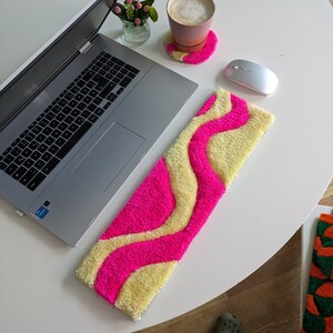 Könnte beinhalten: Ein flauschiger rosa-gelber Teppich mit einem wellenförmigen Muster. Der Teppich liegt auf einem weißen Tisch mit einem Laptop, einer Kaffeetasse und einer Maus.
