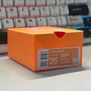 May include: An orange gift box with a white label featuring the text "YOUR NAME", "CLASS OF", and "MAHALO FOR CELEBRATING WITH US". The box has a barcode and the number "26" printed on it. The box is sitting on a desk in front of a keyboard.