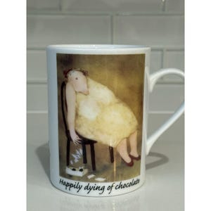 May include: White ceramic mug featuring an illustration of a person slumped in a chair, wearing a yellow dress and red shoes. The mug has the text "Happily dying of chocolate" at the bottom. The mug has a white handle.