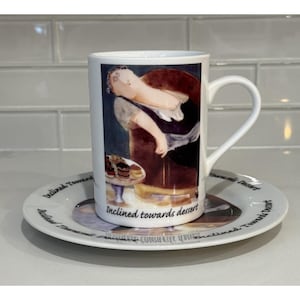 May include: White ceramic mug and saucer set. The mug features a painting of a person reclining, with the words "Inclined towards dessert." The saucer has the same text around the rim. The image is set against a white tile background.