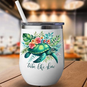 May include: White insulated tumbler with a silver rim and lid, featuring a teal sea turtle design surrounded by flowers and foliage. The tumbler has a white straw and the words "take life slow" in a script font.