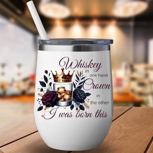May include: A white insulated wine tumbler with a silver rim and lid, featuring the text "Whiskey in one hand, Crown in the other, I was born this." The design includes a whiskey glass, a gold crown, and floral accents.