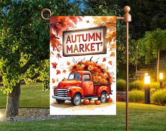 Op de afbeelding: Een tuinvaandel met een rode vintage pick-up truck vol pompoenen en herfstbladeren. De vlag toont de woorden "AUTUMN MARKET" op een houten bord, tegen een achtergrond van herfstbladeren.