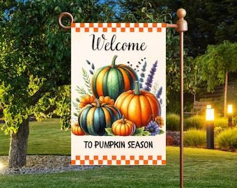 Op de afbeelding: Een tuinvaandel met de tekst "Welcome To Pumpkin Season" en een aquarel illustratie van pompoenen in oranje, groen en blauw. De vlag heeft een oranje en witte geblokte rand.