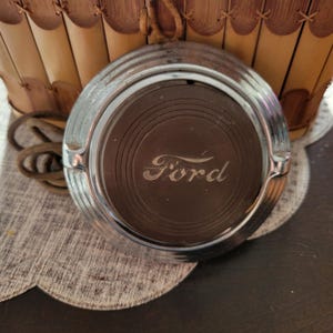 May include: A vintage Ford car emblem with a silver rim and a dark brown center. The word "Ford" is etched in the center. The emblem is resting on a white and beige scalloped doily.