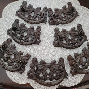 May include: Seven antique bronze drawer pulls with ornate, floral designs. Each pull features a curved handle and decorative flourishes, perfect for adding a vintage touch to furniture. The pulls are arranged on a gray, scalloped placemat.