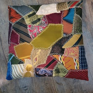 May include: A square throw pillow constructed from a patchwork of various neckties in diverse colors and patterns, including stripes, paisley, and solid textures. The fabric pieces are joined together with visible white decorative stitching.