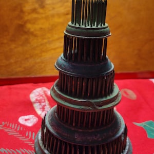 May include: A tiered, dark green and bronze flower frog with numerous metal pins. The flower arranging tool has a circular base and a series of stacked, rounded tiers, each with vertical pins for holding flower stems. The background is a red tablecloth.