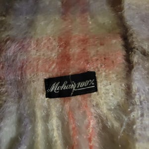 May include: Close-up of a soft, multi-colored mohair scarf. The scarf features a blend of pink, white, and lavender hues. A black label with the text "Mohair 100%" is sewn on the scarf.