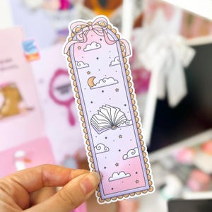 Puede incluir: Marcap&aacute;ginas rectangular con fondo lila claro, con un libro abierto, nubes, estrellas y una luna creciente. El marcap&aacute;ginas tiene un borde festoneado, un lazo rosa y cintas rosas en la parte superior. El dise&ntilde;o es caprichoso y so&ntilde;ador.