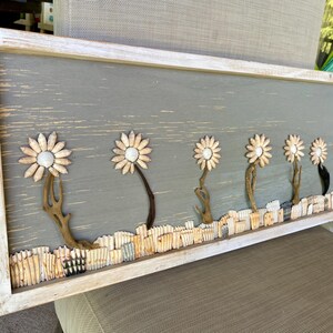 May include: A rectangular wooden wall art piece featuring a row of daisy-like flowers. The flowers have cream-colored petals and white centers, with brown stems. The background is a muted gray, and the frame is white-washed.