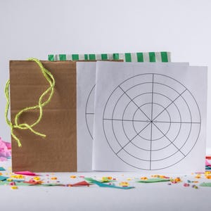 May include: A brown cardboard box tied with yellow twine, with two white sheets of paper featuring a target design. Green and white striped paper and colourful confetti are also present, suggesting a party or game theme.