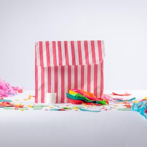 May include: A pink and white striped paper bag with a white roll of tissue paper and colourful streamers inside. The bag is surrounded by colourful tissue paper confetti.