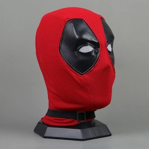 May include: A red Deadpool mask with black eye and mouth details. The mask is made of a textured fabric and features a black strap around the neck. The mask is displayed on a black stand.