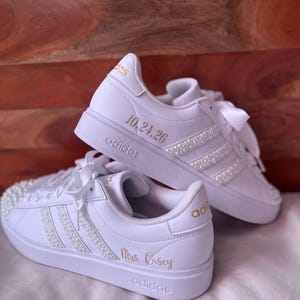 Op de afbeelding: Een paar witte Adidas-sneakers met witte satijnen lintveters en rijen kleine witte parels langs de zijstrepen. De schoenen zijn gepersonaliseerd met gouden letters die 10.24.26 op de hiel en Mrs. Casey aan de zijkant weergeven.
