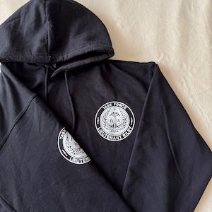 May include: Black hooded sweatshirt with a white embroidered logo on the front and sleeve. The logo is a circle with the text "TASK FORCE 141 LIEUTENANT RILEY 141" around the edge. Inside the circle is a skull with wings and a sword.