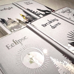 Puede incluir: Una colección de libros con cubiertas plateadas y títulos como "Twilight", "New Moon" y "Breaking Dawn". Cada libro presenta ilustraciones únicas en negro y plata, incluyendo un lobo, árboles y un sol. Los libros están dispuestos de forma ordenada y ligeramente inclinada.