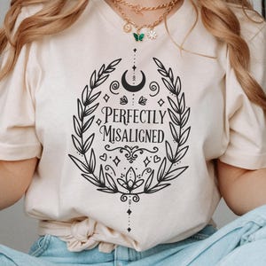 Op de afbeelding: Een crèmekleurig T-shirt met een zwart grafisch ontwerp met een krans van bladeren, een wassende maan en de tekst "Perfectly Misaligned".